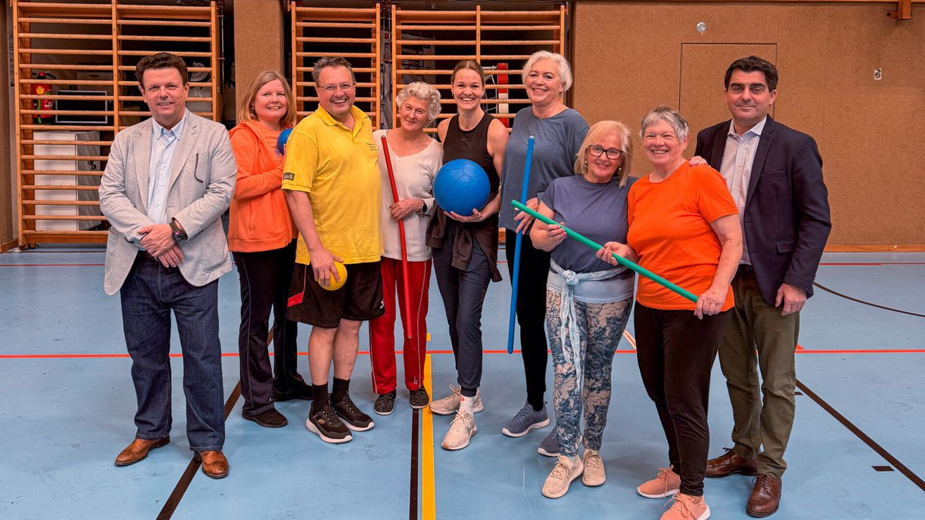 A group of adults, smiling, stand in a gym, holding a blue ball and green sticks. They wear casual clothing.