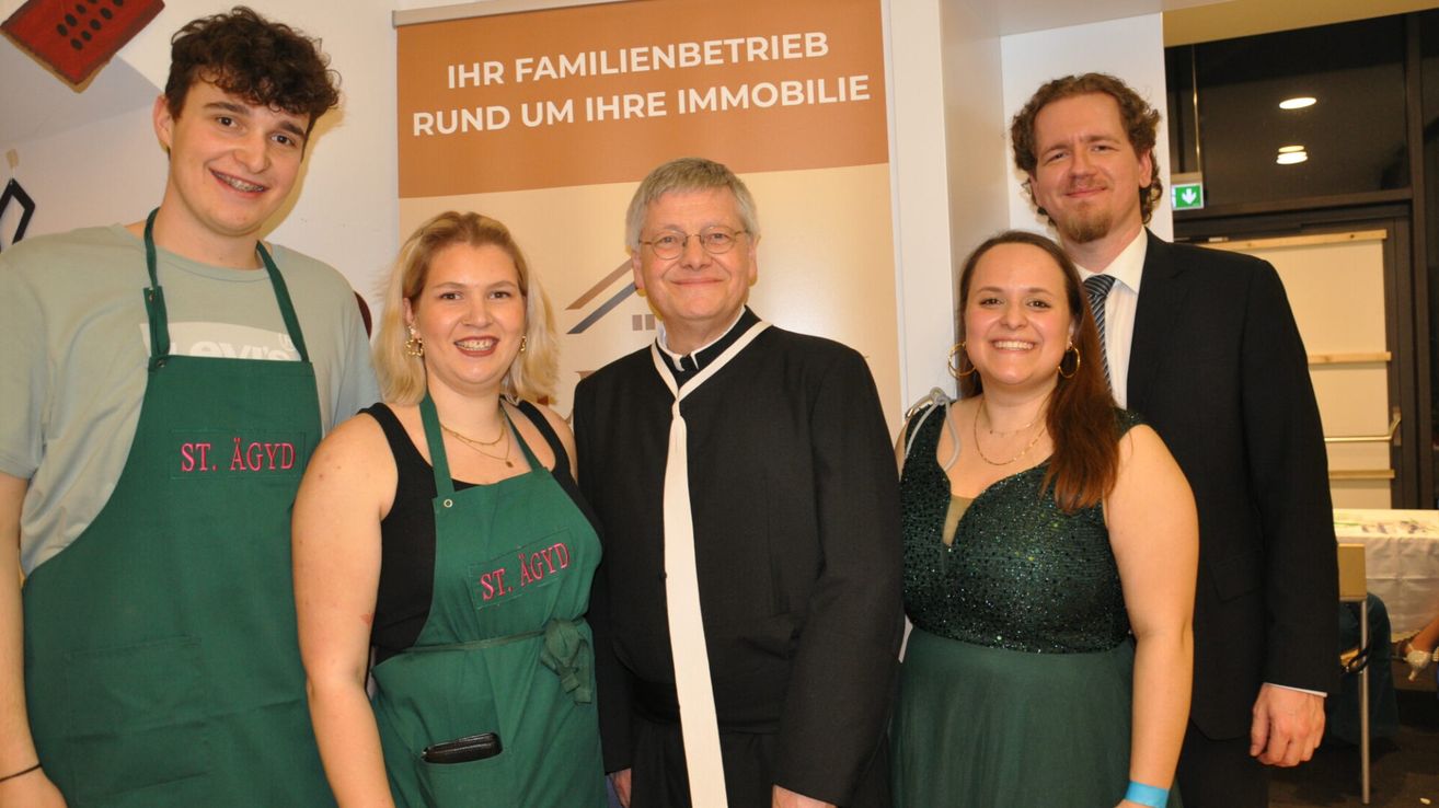 Four people are standing next to each other in front of a sign. The man in the center is wearing glasses. The two women are smiling. The man on the left is wearing a tie.