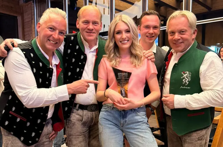 A woman in a pink top holds a Smago Award, smiling. She is surrounded by four men in traditional clothing. They are all smiling and looking at the camera.