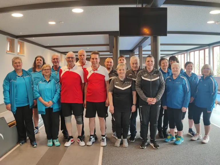 A group of people, possibly athletes, stands in a line wearing sports attire, smiling and posing for a photo. The setting appears to be an indoor sports facility.