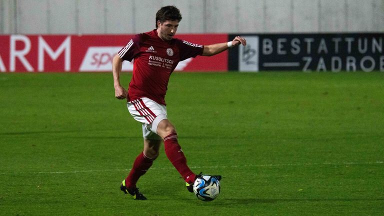 A soccer player in a maroon jersey kicks a ball on a field. The player wears red and white shorts, red socks, and black cleats.
