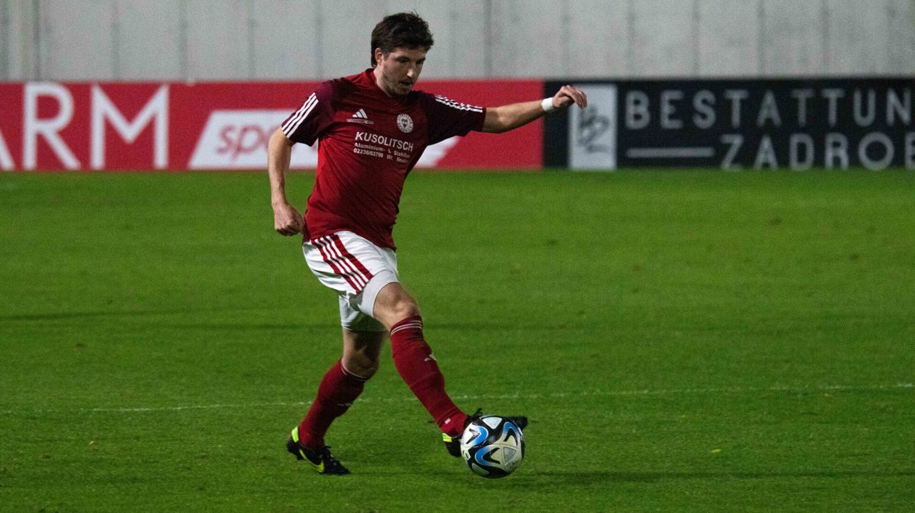 A soccer player in a maroon jersey kicks a ball on a field. The player wears red and white shorts, red socks, and black cleats.