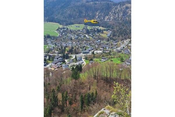 A yellow helicopter flies over a village with many buildings and trees. The village is surrounded by mountains and green fields.