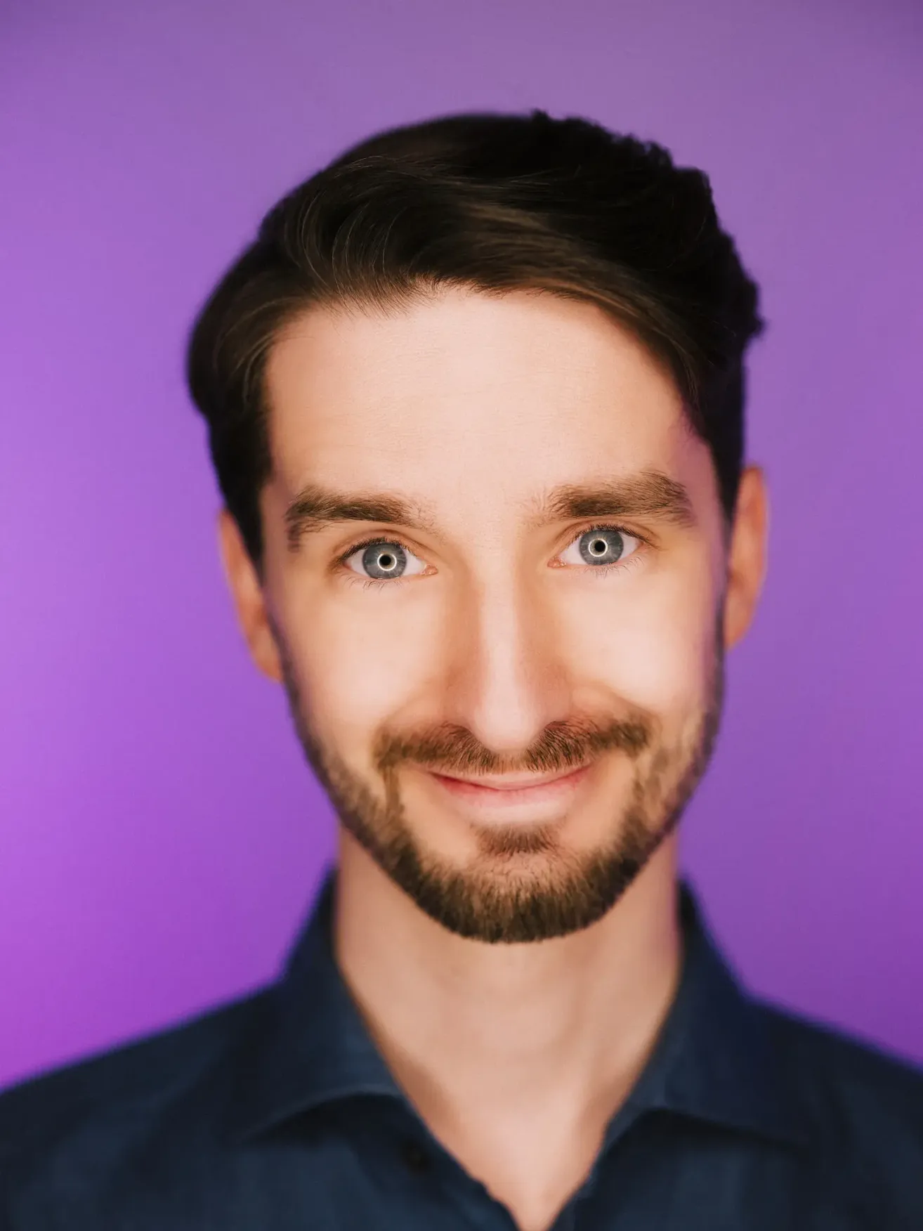 Ein lächelnder Mann mit Bart und blauen Augen, in einem dunkelblauen Hemd, vor einem lila Hintergrund.