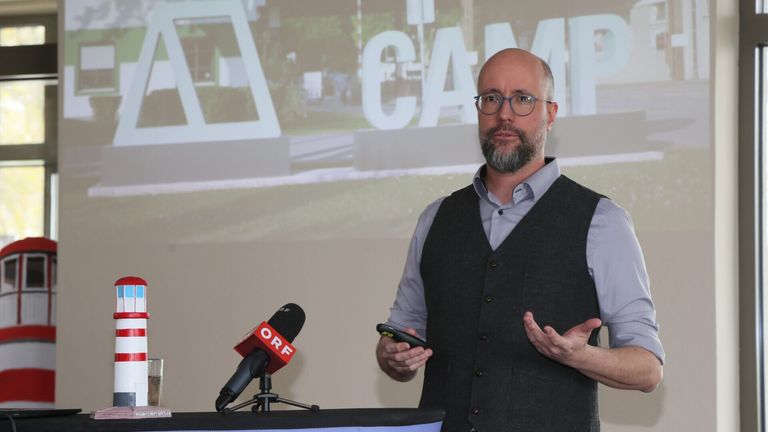 A man stands at a podium with a microphone, speaking to an audience. Behind him, a large screen displays the word 'CAM'. He is dressed in a vest and holds a remote control.