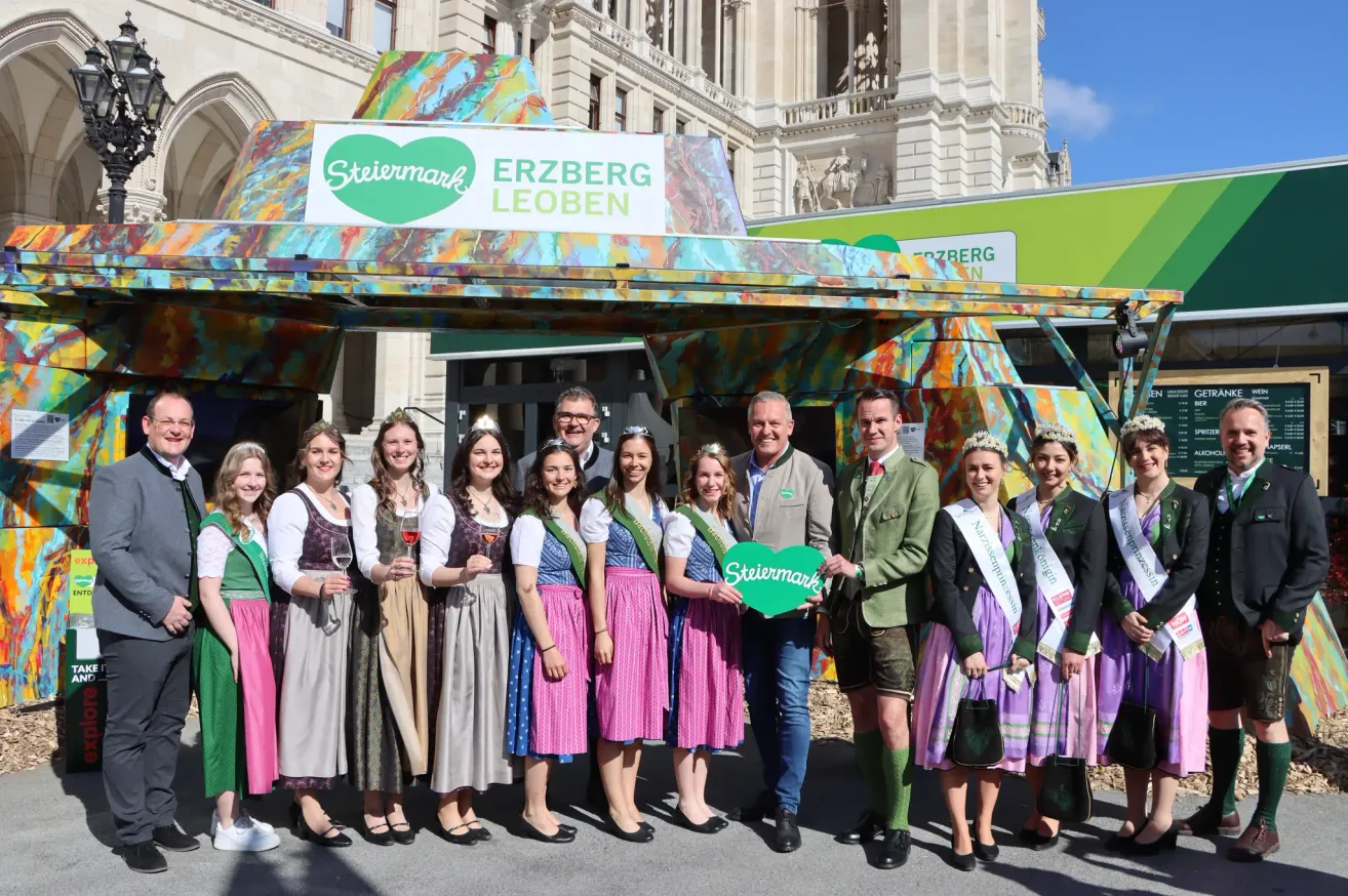 Eine Gruppe von Menschen in traditioneller Kleidung, die vor einer farbenfrohen Erzberg-Veranstaltungsstruktur stehen. Dahinter befindet sich ein Gebäude mit Statuen, und sie halten ein grünes Schild mit der Aufschrift 'Stiermark'.