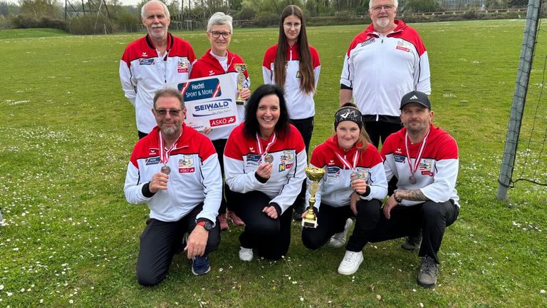 A group of people, likely athletes, pose for a photo on a grassy field. They wear red and white jackets, with some holding trophies and medals. A woman holds a sign with the word 'Sport'. The background shows trees and a fence.