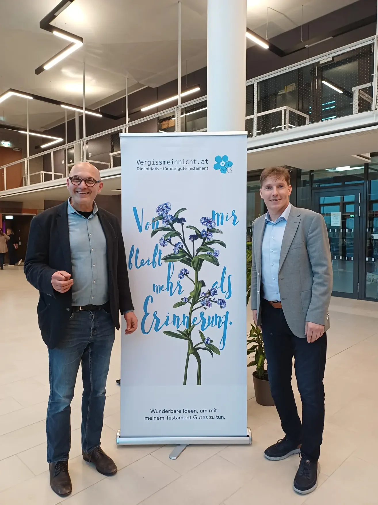 Two men are standing in front of a white banner with a floral design, smiling for the camera. The banner reads 'Vergissmeinnicht' and 'mir mehr Erinnerung' in blue font.