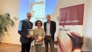 Drei Personen stehen lächelnd und halten Bücher in einer Hand. Eine Projektionsleinwand zeigt ein Buchcover. Der Banner liest 'LESEN LIEBEN' und eine Website.