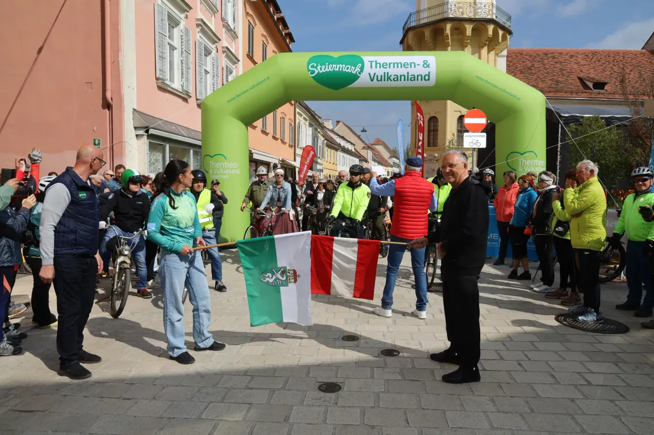 Eine Gruppe von Radfahrern versammelt sich unter einem Torbogen, ein Mann hält Flaggen und ein anderer Mann steht neben ihnen, während andere Helme und Jacken tragen. Gebäude mit Vordächern sind hinter ihnen.