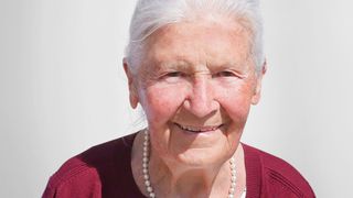 An elderly woman with white hair, wearing a red sweater and pearl necklace, smiling at the camera.