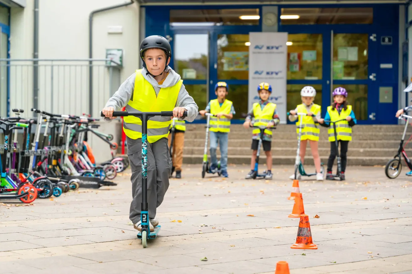 A young boy in a yellow vest rides a scooter through an obstacle course with other children, in front of a building.
