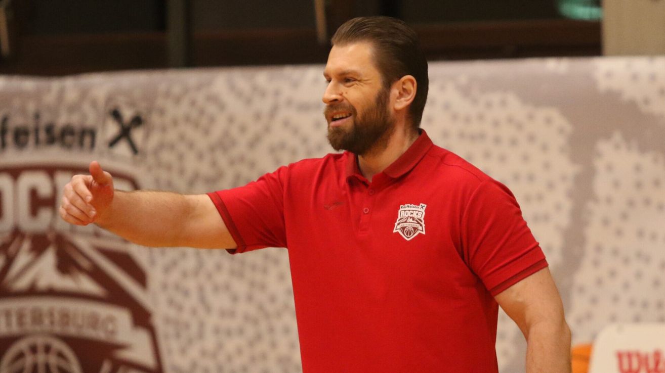 A man with a beard in a red polo shirt is smiling and pointing in a basketball court.