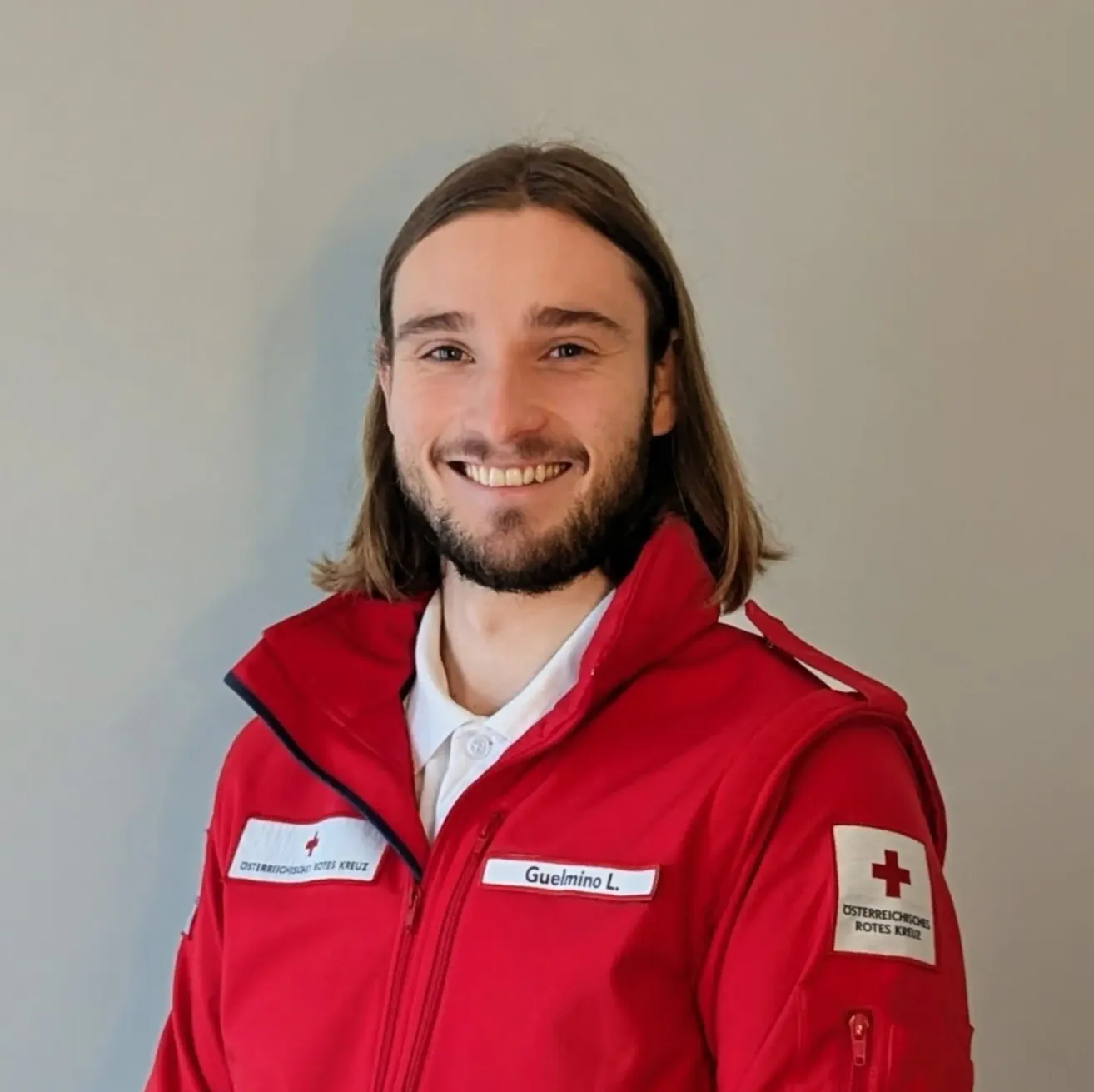 A man in a red jacket with a white collar stands smiling, with a red cross on his shoulder. His name is Guelimino L.