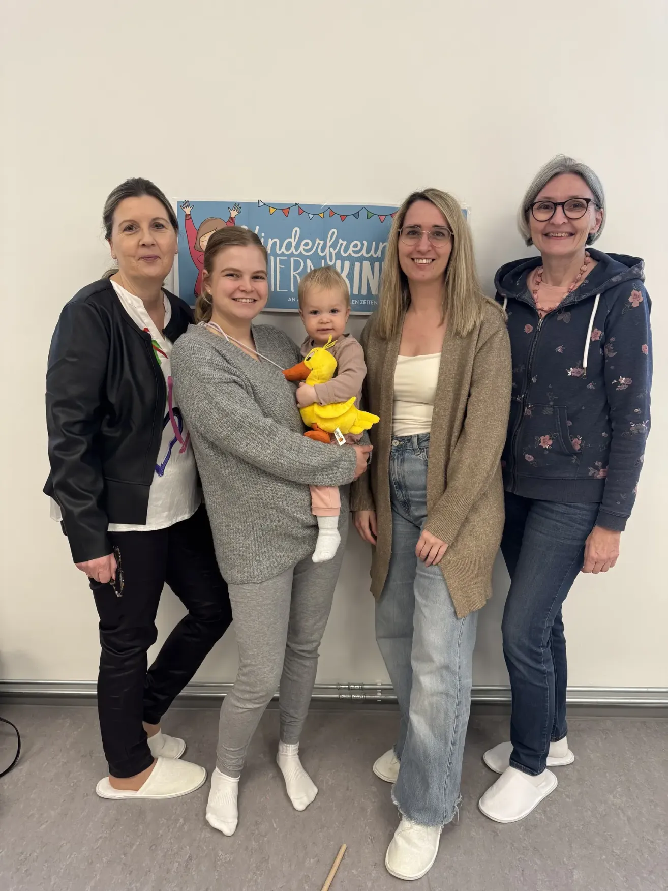 Four women and a baby pose for a photo in a room with a white wall and a banner reading Kinderfreunde. The woman on the left holds a baby, while the others stand beside her smiling.