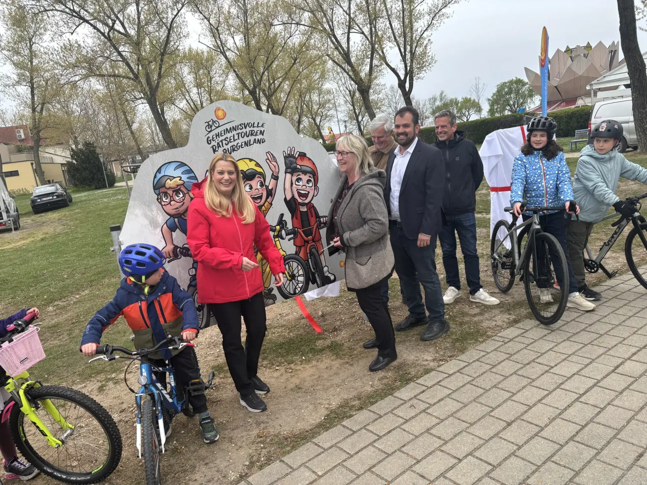 Eine Gruppe von Menschen steht vor einem Schild mit Comicfiguren und Fahrrädern und lächelt. Hinter ihnen fahren ein Junge und ein Mädchen auf einem gepflasterten Weg mit Fahrrädern.