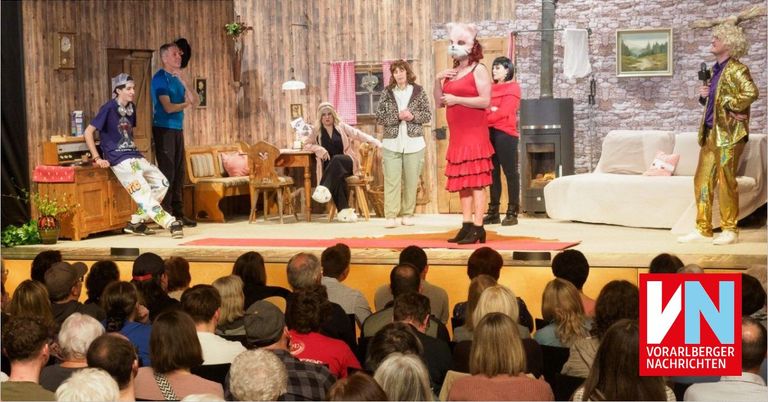 A stage performance with a group of actors and a masked woman. Behind them, a cozy setting with furniture and a fireplace. The audience is seated and watching.