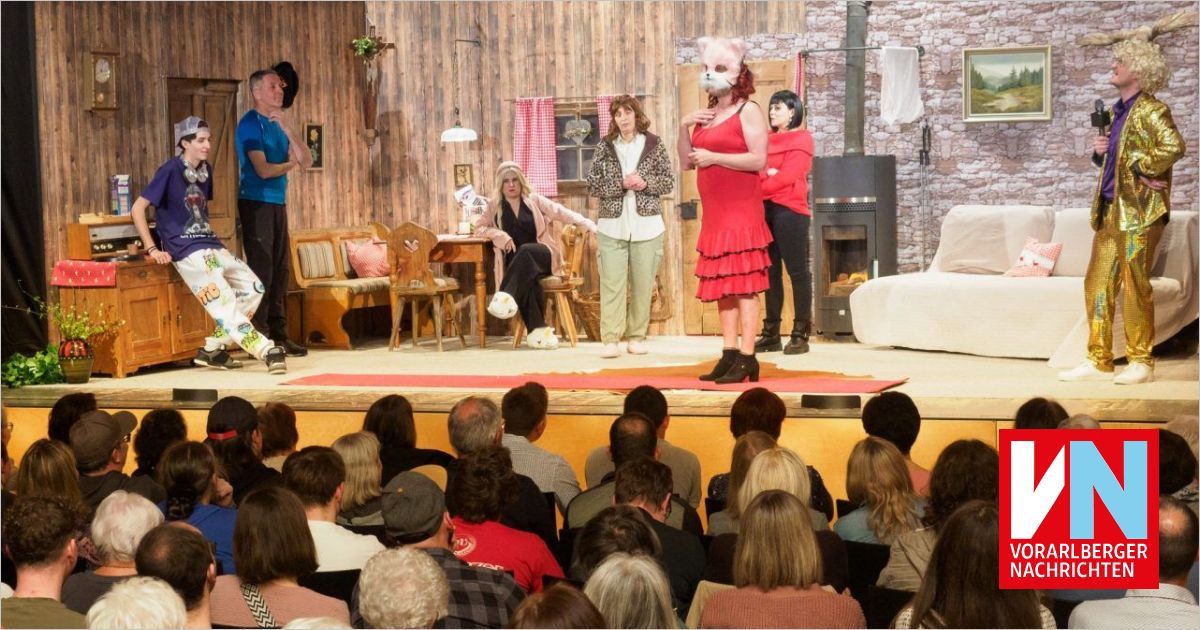 A stage performance with a group of actors and a masked woman. Behind them, a cozy setting with furniture and a fireplace. The audience is seated and watching.