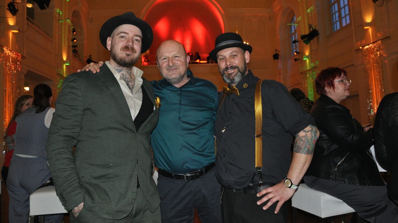 Three men stand close together, smiling for a photo. The left man wears a hat and a green suit. The middle man has a beard and wears a blue shirt. The right man has a bow tie and suspenders, wearing a black shirt. Behind them is a red-lit room with a balcony.