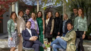 A group of people stand and sit on chairs in front of a modern building with glass walls, surrounded by trees and greenery. A man sits on a chair with a green table, holding flowers.