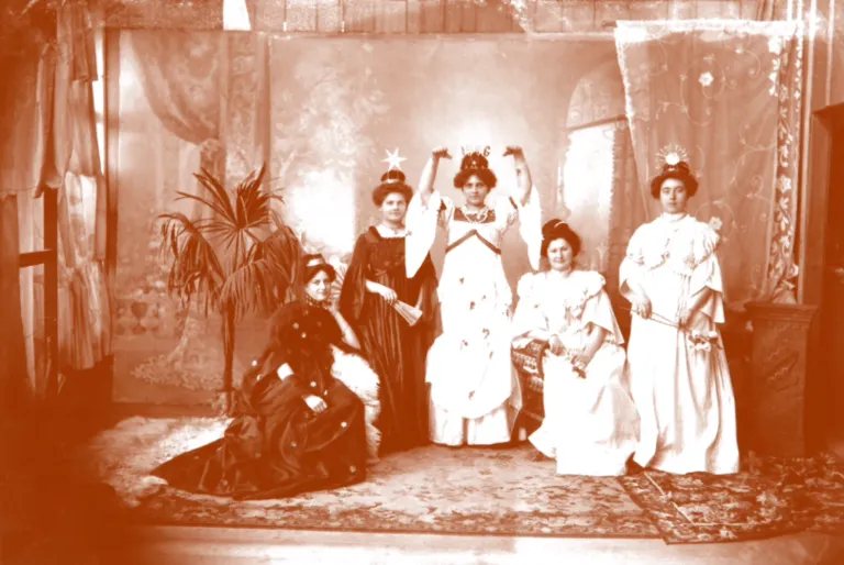 A vintage photo shows five women dressed in white, posing. Three stand with raised hands, while two sit. One holds a fan, and all wear hats. A potted plant is on the left.