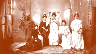 A vintage photo shows five women dressed in white, posing. Three stand with raised hands, while two sit. One holds a fan, and all wear hats. A potted plant is on the left.