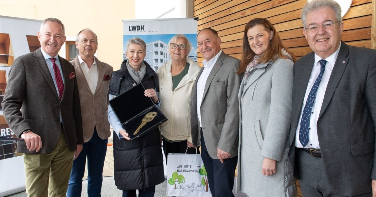 A group of people, including women and men, are standing together outside. One woman holds a key and a box, while a man holds a bag. They are all smiling and standing near a wooden wall with a sign.