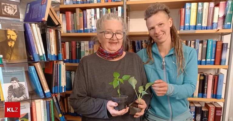 Two women in a library, one with glasses and a scarf holds plants, the other in a blue jacket smiles.