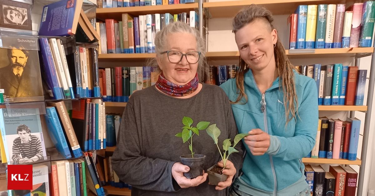 Two women in a library, one with glasses and a scarf holds plants, the other in a blue jacket smiles.