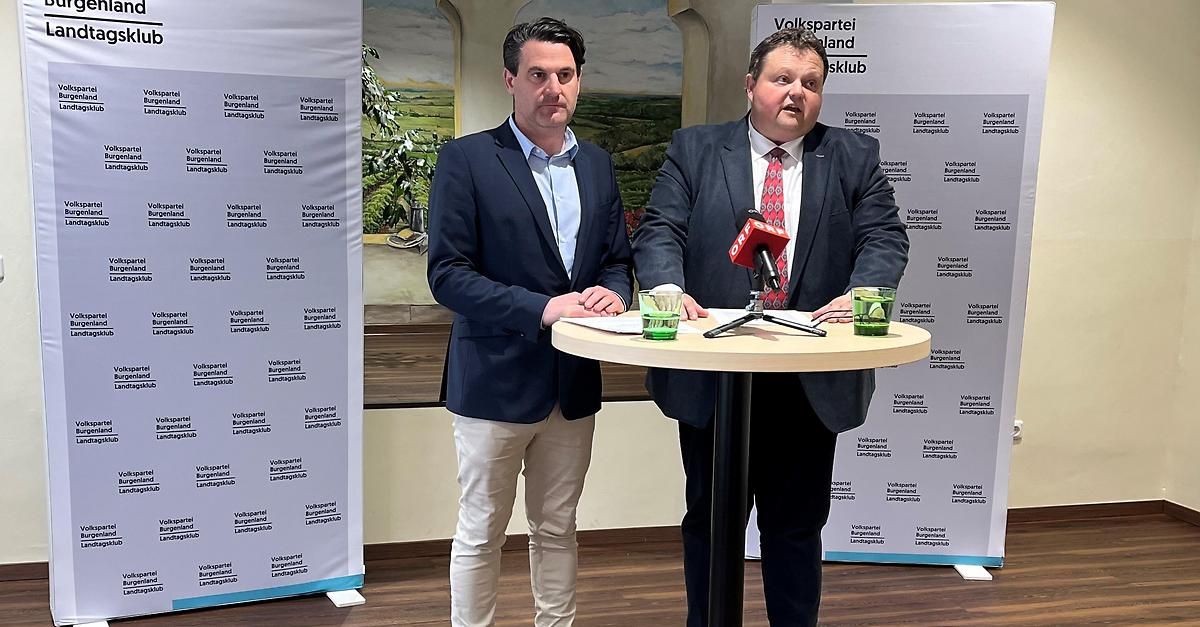 Two men stand behind a table with microphones at a press conference. Both wear suits and are speaking to the camera.