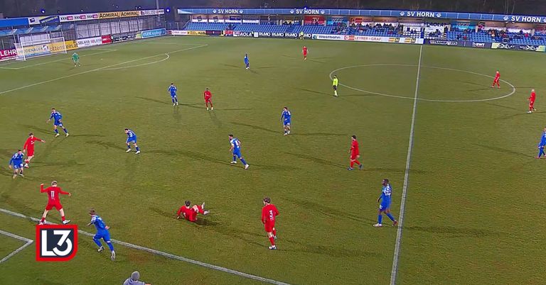 Soccer players in blue and red uniforms are on a field. The red team is attacking, while the blue team defends. The referee stands near the center circle.