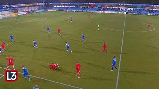 Fußballspieler in blauen und roten Uniformen sind auf einem Feld. Die rote Mannschaft greift an, während die blaue Mannschaft verteidigt. Der Schiedsrichter steht in der Nähe des Mittelkreises.