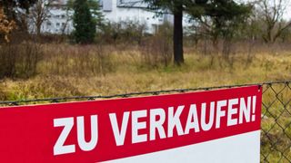 A red and white sign with white letters saying 'ZU VERKAUF' on it, in front of a field and a white house.