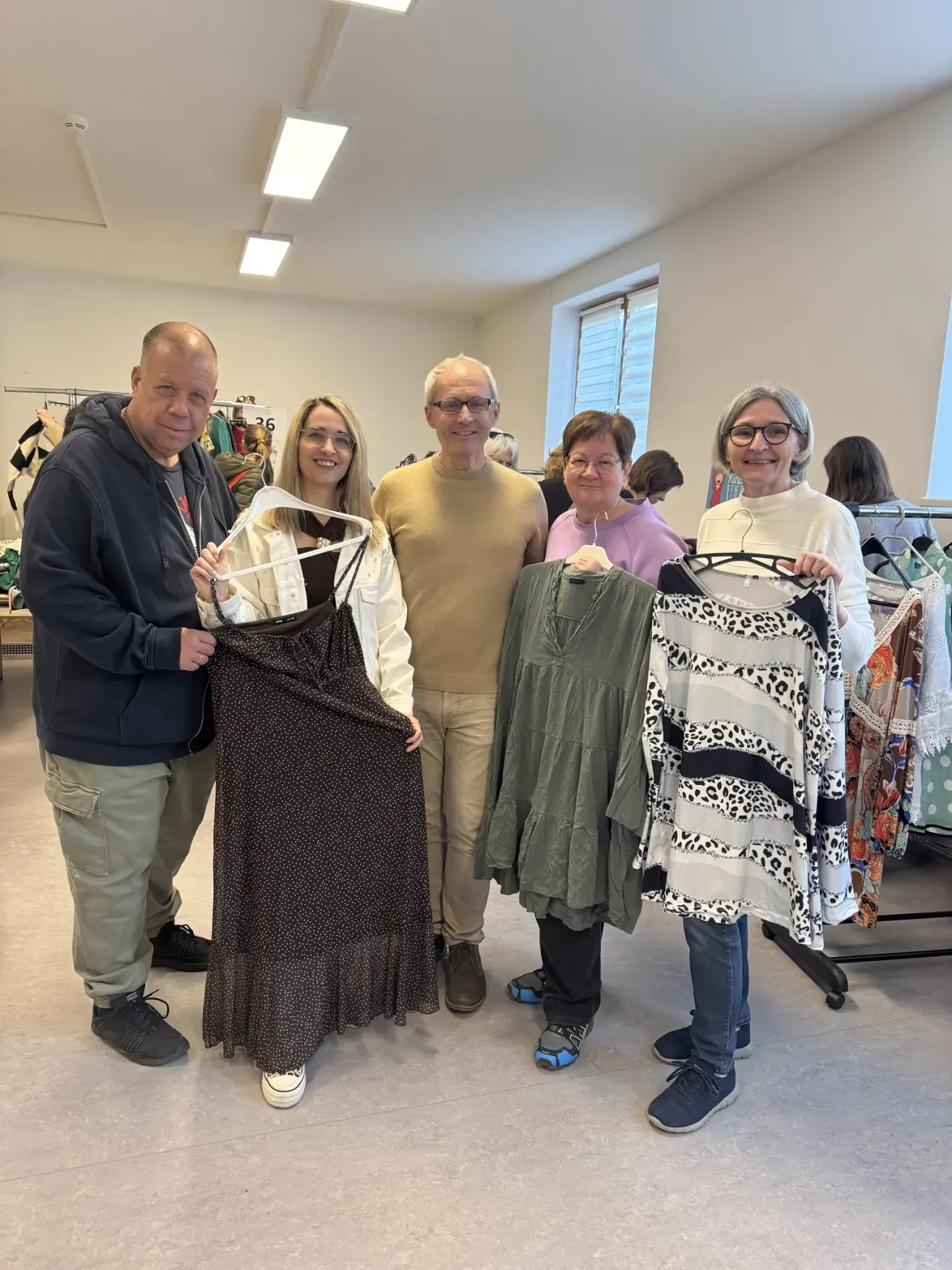 Five adults smiling in a clothing store. Three women hold up dresses, and two men stand next to them.