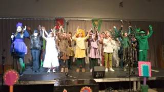 A group of children in costumes perform on stage with microphones and speakers in the background.