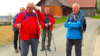 Four older men in hiking gear are standing on a pathway. One man has a red jacket, another blue. They are smiling and appear to be discussing something. A barn is visible in the background.