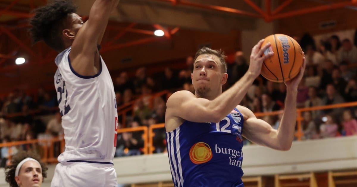 A basketball player in a blue jersey shoots the ball. An opponent in white tries to block the shot. They are in a crowded arena.