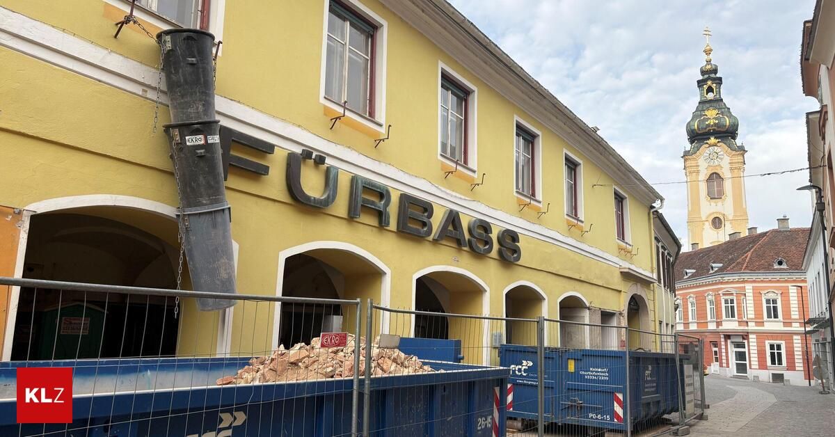 A construction site with a yellow building, 'Furbass' on its sign. Two dumpsters are filled with rubble, fenced off.