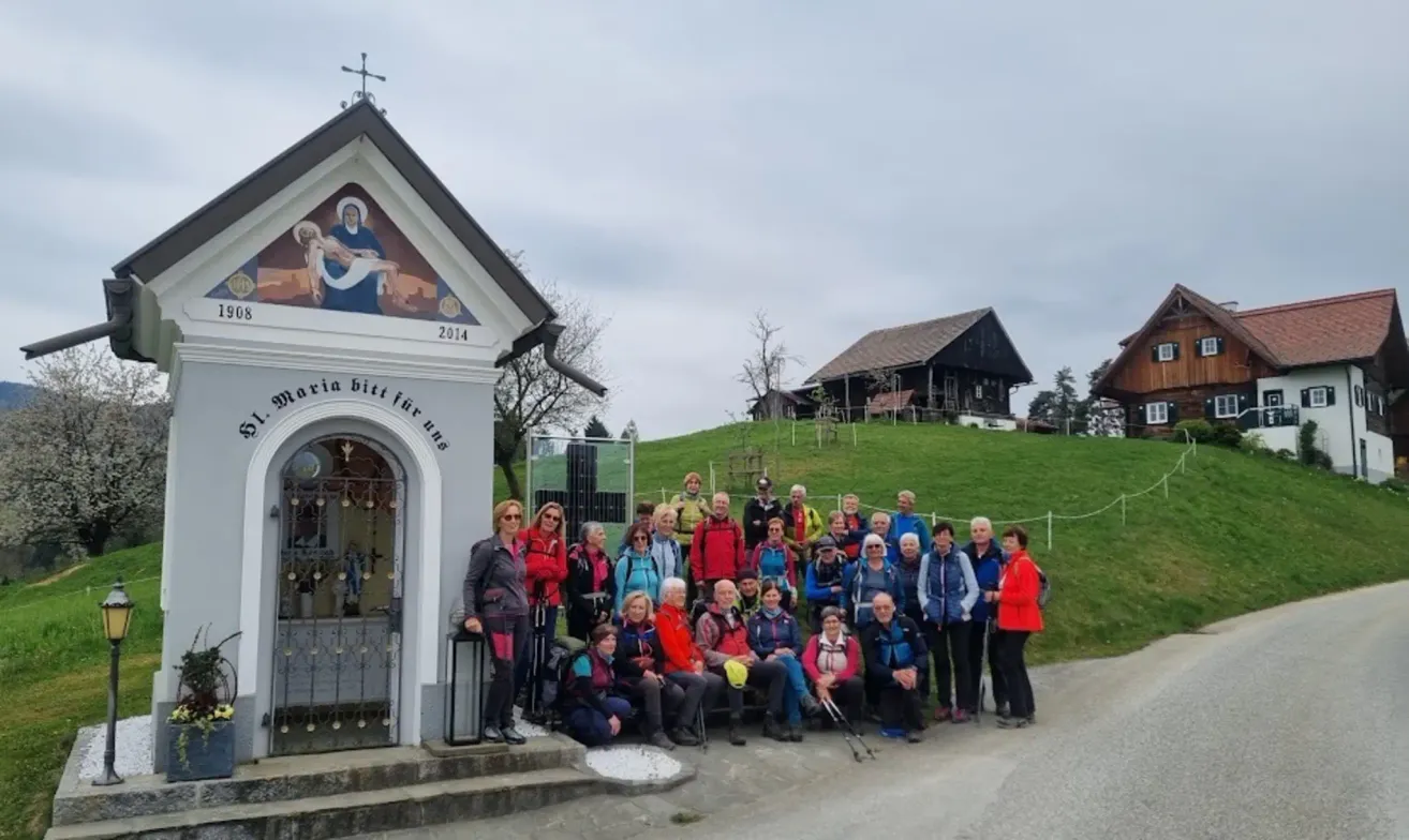 A group of hikers poses for a photo in front of a chapel in a rural area, with a house and green hill in the background.