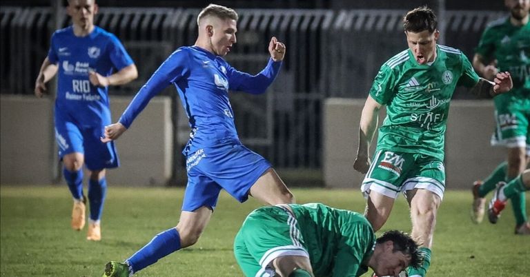 A soccer player in blue slides on the field, while a player in green falls to the ground, and another player in green stands behind him.