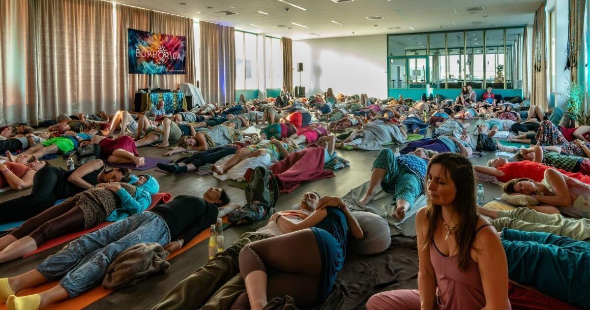 Viele Menschen liegen auf Yogamatten, einige in bunten Kleidern. Ein Banner mit dem Text 'Euphoria' hängt an der Wand. Menschen sitzen oder liegen herum, manche ruhen sich aus.