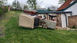 A red tractor is overturned on a grassy lawn, with its front facing upward and a wheel detached. Behind it, there's a wooden fence and a building with a black roof.