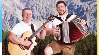 Two men stand in front of mountains, one with a guitar, the other with an accordion. Both are smiling, dressed in traditional Alpine attire.