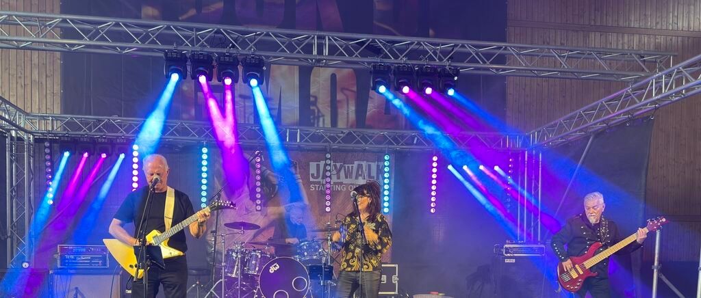 Eine Band tritt auf einer Bühne mit leuchtenden lila und blauen Lichtern auf. Ein Mann spielt Gitarre und singt in ein Mikrofon.