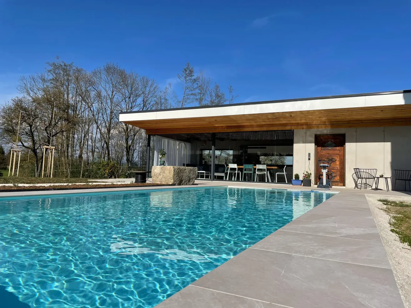 Ein modernes Haus mit einem großen Swimmingpool davor, umgeben von Bäumen und mit einer Holzüberdachung und Glaswänden.