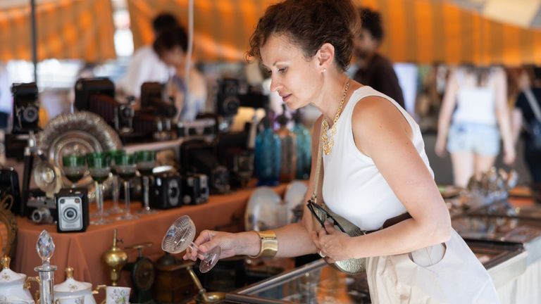 A woman examines a clear glass wine glass at a flea market, surrounded by various items like cameras and glassware.