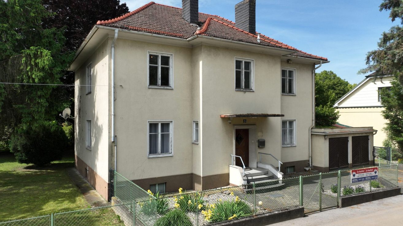 Ein zweistöckiges beiges Haus mit braunem Rand. Es gibt zwei Schornsteine auf dem Dach, drei Fenster auf der linken Seite und zwei Fenster auf der rechten Seite. Vor dem Haus befindet sich ein kleiner Garten.