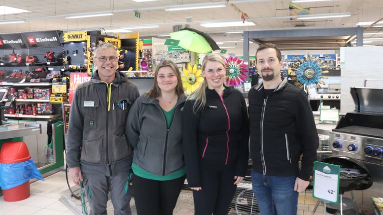 Four people stand in a store with a smile, displaying various items in the background. A man on the left wears glasses, while a woman next to him has a necklace. Another woman wears glasses and a jacket with a pink stripe. A man on the right has a beard.