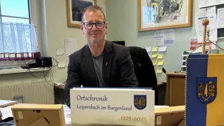 A man is smiling in an office, wearing glasses and a suit. A book titled Ortschronik Loipersdorf im Burgenland 1225-2025 is on his desk. Two framed pictures and sticky notes are on the wall.