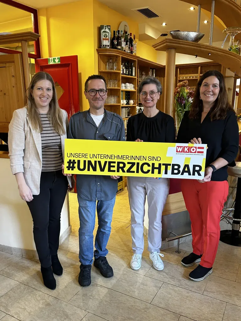 Vier Personen stehen zusammen und halten ein gelbes Banner mit der Aufschrift Unsere Unternehmer:innen sind #unverzichtbar. Sie lächeln und posieren für ein Foto in einem Restaurant.